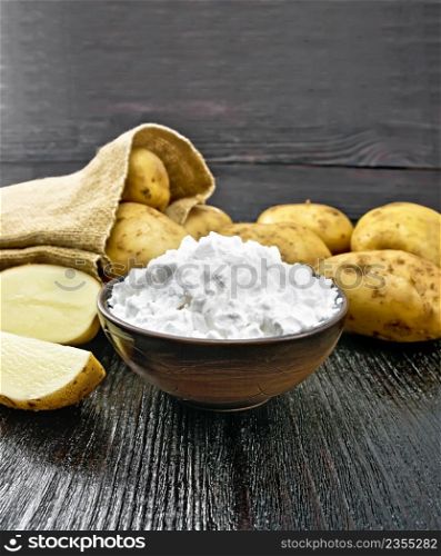 Potato starch in clay bowl, vegetable tubers in a bag and on the table against the background of black wooden board