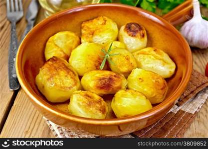 Potatoes roasted with rosemary in a ceramic pan on a napkin, garlic, parsley, vegetable oil on a wooden boards background