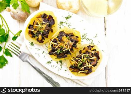 Potatoes stuffed with mushrooms, fried onions and cheese in a plate on napkin, vegetable oil in decanter, parsley, garlic and a fork on the background of light wooden board from above