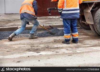 Pothole repair of the road. A team of workers patches holes in the old pavement with fresh asphalt.. A road maintenance worker distributes fresh asphalt with a shovel at the patching of the road.