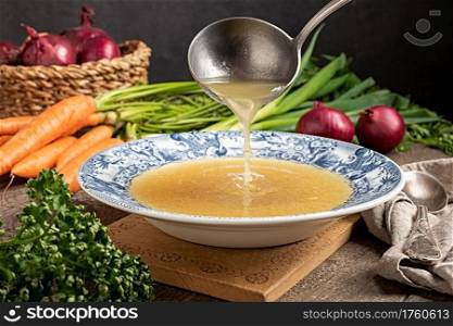 Pouring chicken bone broth from a ladle into a vintage plate, with fresh vegetables in the background