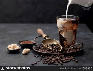Pouring fresh milk into glass of iced black coffee on tray with beans and ground coffee with cane sugar and vintage shop on black background. Space for text