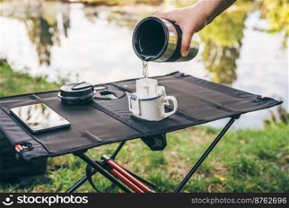 Pouring hot water through trendy convenient paper drip coffee bag into metal cup on c&ing table outdoors. Making freshly brewed coffee in nature. Pouring hot water through drip coffee bag outdoors