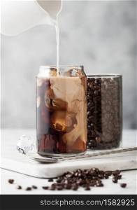 Pouring milk into glass with iced black coffee with glass container of beans and spoon on marbel board and light table background.