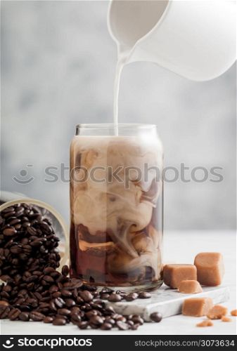 Pouring milk into glass with iced black coffee with glass jar of coffee beans and salted caramel on light table background.