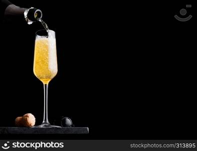 Pouring yellow champagne from bottle to glass with cork and wire cage on black marble board on black background.