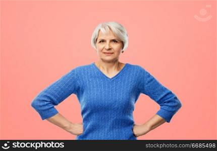 power and old people concept - portrait of senior woman in blue sweater with hands on hips over pink or living coral background. senior woman in blue sweater hands on hips