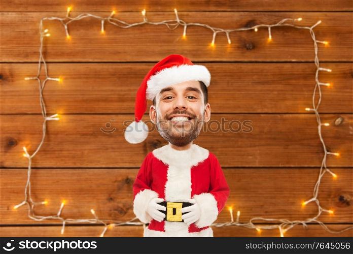 power, fitness, strength, sport and people concept - happy smiling young man in costume of santa over garland on wooden background (funny cartoon style character with big head). happy santa over garland on wooden background