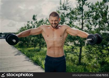 Powerful muscular sportsman demonstrates endurance, lifts heavy barbells and trains arms muscles, poses shirtless outdoor, puts all efforts in lifting weight. Athletic man works out with dumbbells