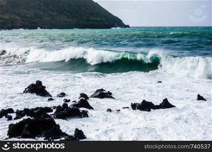 Powerful ocean waves crashing on jagged basalt rocks with island in background, in Azores, Portugal.