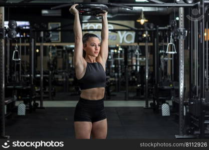 Powerful young fit female athlete in sports top and shorts holding heavy weight plate over head and looking away while doing press exercise in modern gym. Strong confident sportswoman doing overhead press with weight plate