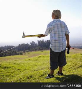 Pre-teen Caucasin male holding remote control and airplane standing on a grassy knoll.