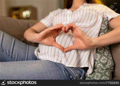 pregnancy and people concept - close up of pregnant woman on sofa showing hand heart gesture at home. pregnant woman showing hand heart gesture