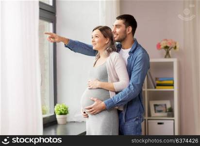 pregnancy and people concept - happy man hugging his pregnant wife and showing something outside window at home. man showing something out window to pregnant wife