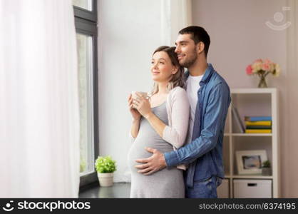 pregnancy and people concept - happy man hugging his pregnant wife standing at window at home. man hugging pregnant woman at window at home