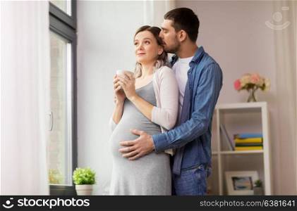 pregnancy and people concept - happy man hugging his pregnant wife standing at window at home. man hugging pregnant woman at window at home