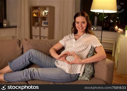 pregnancy and people concept - happy smiling pregnant woman on sofa showing hand heart gesture at home. happy pregnant woman showing hand heart gesture