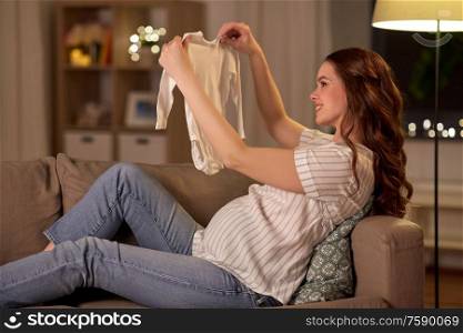pregnancy and people concept - happy smiling pregnant woman with baby bodysuit on sofa at home. happy pregnant woman with baby bodysuit at home