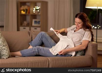 pregnancy and people concept - happy smiling pregnant woman with baby bodysuit on sofa at home. happy pregnant woman with baby bodysuit at home