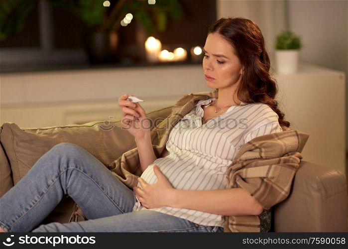 pregnancy and people concept - sad sick pregnant woman measuring temperature with thermometer on sofa at home. sick pregnant woman with thermometer at home