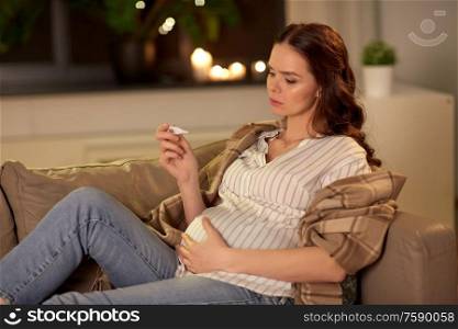 pregnancy and people concept - sad sick pregnant woman measuring temperature with thermometer on sofa at home. sick pregnant woman with thermometer at home