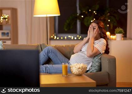 pregnancy and people concept - scared pregnant woman on sofa watching tv at home. scared pregnant woman watching tv at home