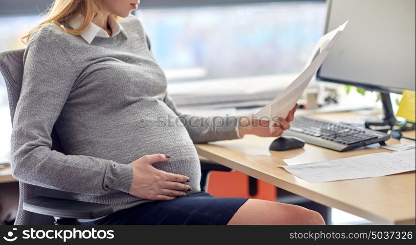 pregnancy, business and work concept - smiling pregnant businesswoman sitting at office table and reading papers. pregnant businesswoman reading papers at office