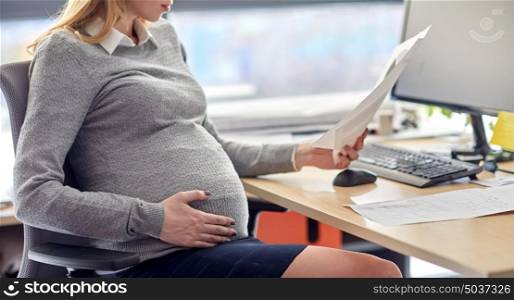 pregnancy, business and work concept - smiling pregnant businesswoman sitting at office table and reading papers. pregnant businesswoman reading papers at office