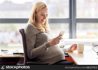 pregnancy, business, work and technology concept - smiling pregnant businesswoman with smartphone and tea cup sitting at office table