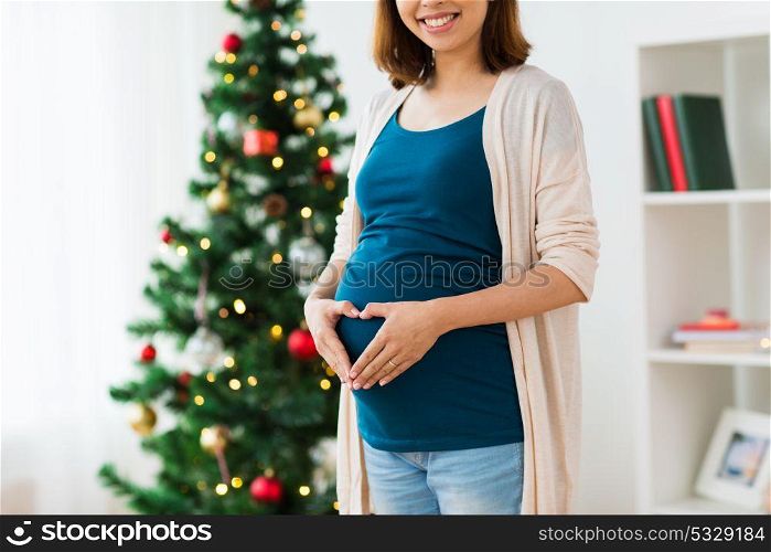 pregnancy, christmas and people concept - close up of pregnant woman making hand heart on belly at home. close up of pregnant woman with heart at christmas