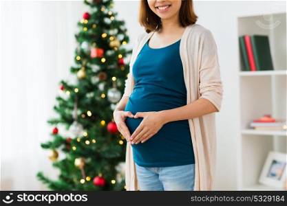 pregnancy, christmas and people concept - close up of pregnant woman making hand heart on belly at home. close up of pregnant woman with heart at christmas