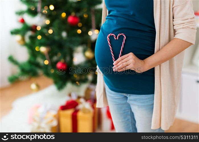 pregnancy, christmas and people concept - close up of pregnant woman with heart made of candy canes on belly at home. close up of pregnant woman with heart at christmas
