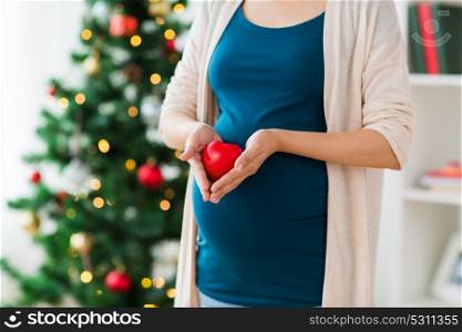 pregnancy, christmas and people concept - close up of pregnant woman with red heart on belly at home. close up of pregnant woman with heart at christmas