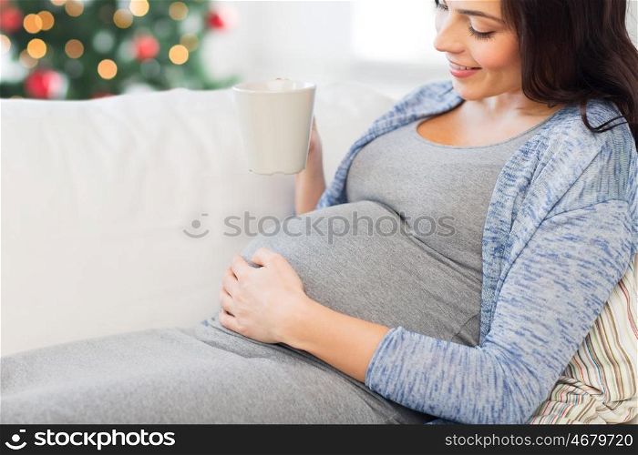 pregnancy, drinks, holidays, people and expectation concept - close up of happy pregnant woman with big belly holding cup and drinking tea over christmas tree background