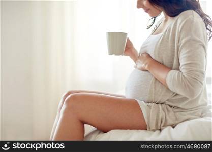 pregnancy, drinks, rest, people and expectation concept - close up of happy pregnant woman with cup drinking tea sitting on bed at home bedroom