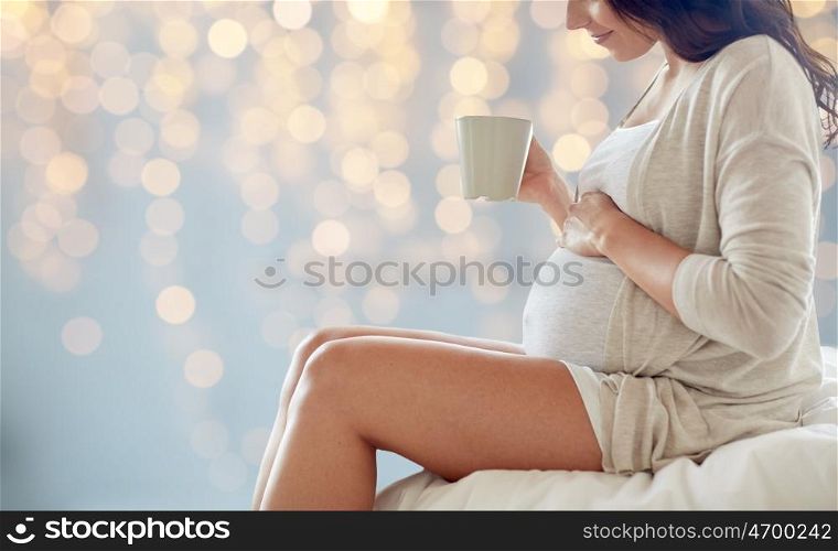 pregnancy, drinks, rest, people and expectation concept - close up of happy pregnant woman with cup drinking tea sitting on bed over holidays lights background