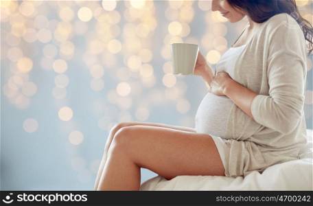 pregnancy, drinks, rest, people and expectation concept - close up of happy pregnant woman with cup drinking tea sitting on bed over holidays lights background