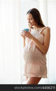pregnancy, drinks, rest, people and expectation concept - happy pregnant woman in nightie drinking tea from cup at home