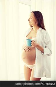 pregnancy, drinks, rest, people and expectation concept - happy pregnant woman in shirt and underware with cup drinking tea at home