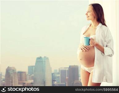 pregnancy, drinks, rest, people and expectation concept - happy pregnant woman in shirt and underwear with cup drinking tea over city background