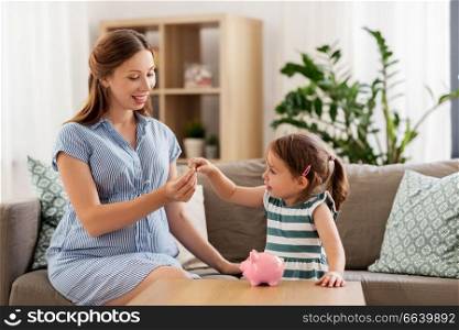 pregnancy, finances and family concept - happy pregnant mother and little daughter putting euro coin into piggy bank at home. pregnant mother and daughter with piggy bank