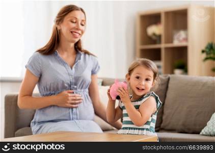 pregnancy, finances and family concept - happy pregnant mother and little daughter with piggy bank at home. pregnant mother and daughter with piggy bank