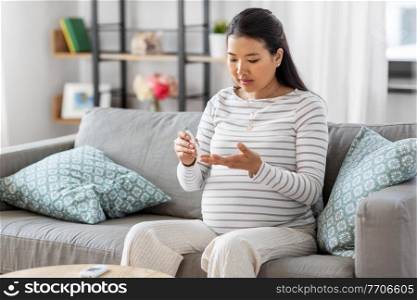 pregnancy, health and glycemia concept - pregnant asian woman checking blood sugar level with glucometer and lancing device at home. pregnant woman with glucometer making blood test
