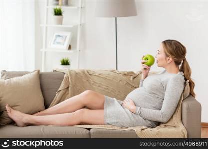 pregnancy, health, food and people concept - happy pregnant woman sitting on sofa and eating green apple. happy pregnant woman eating green apple