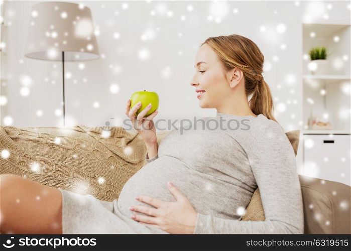 pregnancy, health, food and people concept - happy pregnant woman sitting on sofa with green apple over snow. happy pregnant woman with green apple