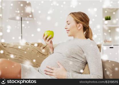 pregnancy, health, food and people concept - happy pregnant woman sitting on sofa with green apple over snow. happy pregnant woman with green apple