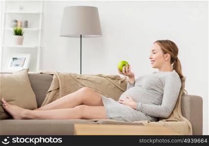 pregnancy, health, food and people concept - happy pregnant woman sitting on sofa with green apple. happy pregnant woman with green apple