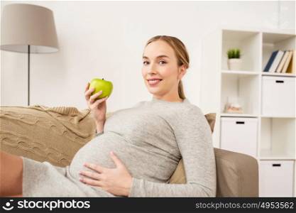 pregnancy, health, food and people concept - happy pregnant woman sitting on sofa with green apple. happy pregnant woman with green apple