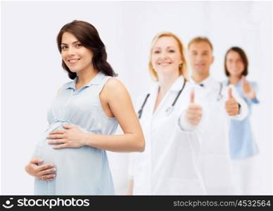 pregnancy, healthcare, medicine, people and expectation concept - happy pregnant woman touching her big belly over group of doctors or obstetricians showing thumbs up background