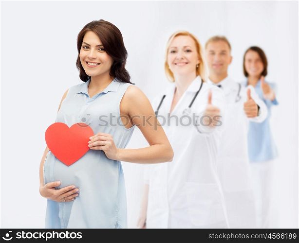 pregnancy, healthcare, medicine, people and expectation concept - happy pregnant woman with red heart shape touching her belly over group of doctors or obstetricians showing thumbs up background